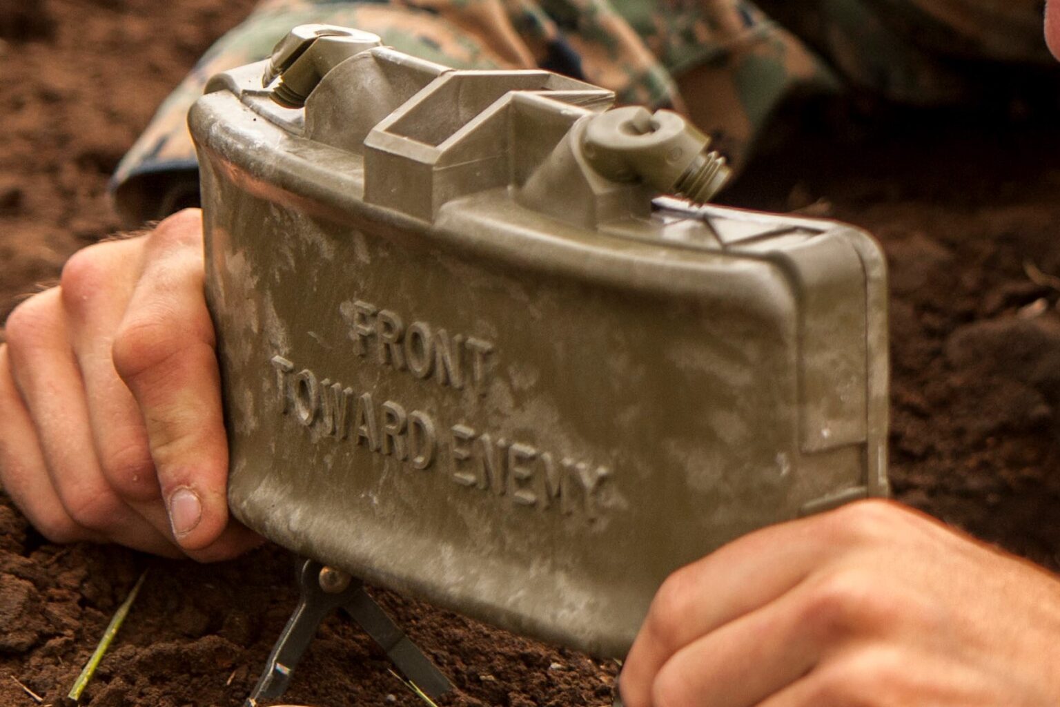 Person holding a Claymore mine device with 'Front Toward Enemy' text, on a dirt background.